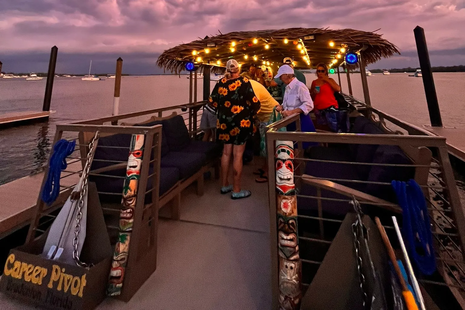 On deck at dusk under the thatched roof and string lights, carved tiki posts either side, a pink sky overhead
